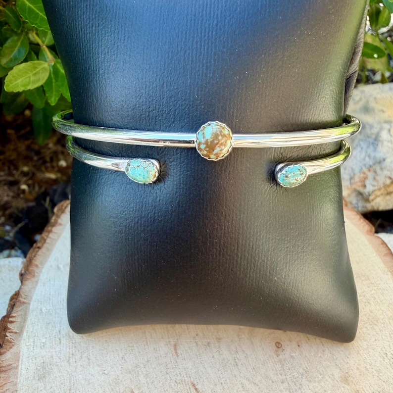 May include: A silver-toned cuff bracelet with three turquoise and brown stones. The bracelet has a simple, open design with a central round stone and two oval stones on the ends. The bracelet is displayed on a black surface.
