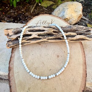 May include: White beaded necklace with silver accents. The necklace is displayed on a wooden surface with the handwritten text "Ally k". The beads are small and evenly spaced, creating a delicate and elegant look. The necklace is a simple yet stylish accessory.