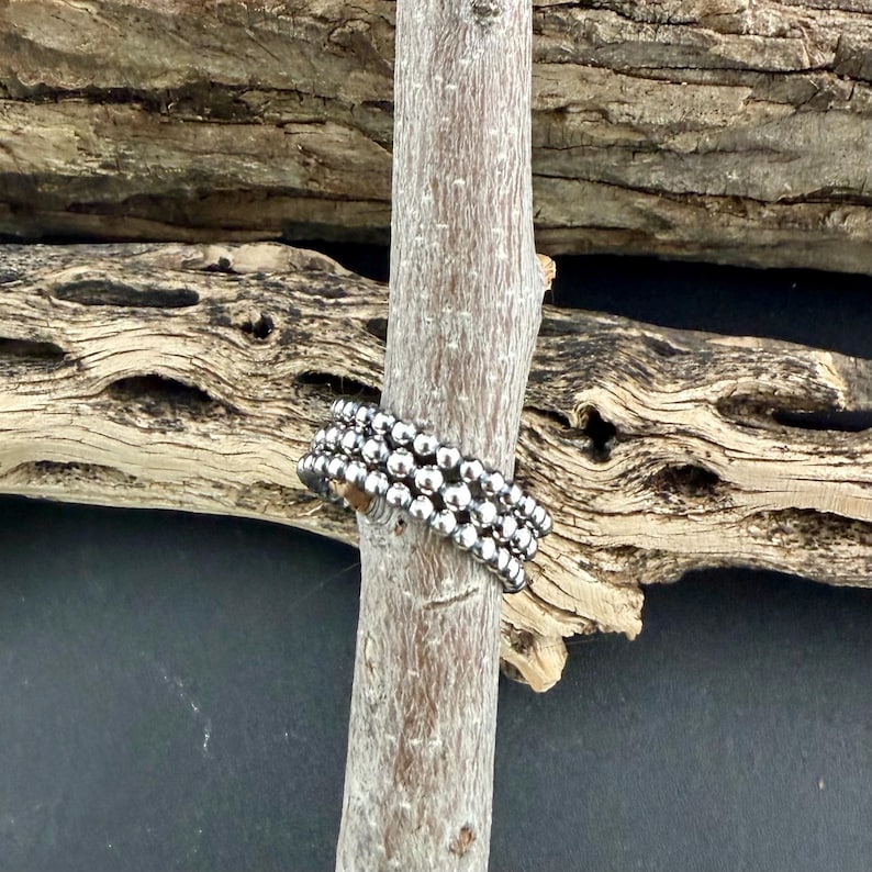Puede incluir: Un anillo plateado con cuentas con tres filas de peque&ntilde;as cuentas redondas. El anillo se muestra en un palo de madera marr&oacute;n claro, con un fondo de madera texturizada. Una joya sencilla pero elegante.