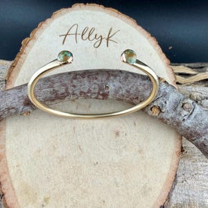 May include: A gold-colored cuff bracelet with turquoise-colored stones at each end. The bracelet is displayed on a wooden slice with the name "Allyk" engraved on it. The background includes a wooden branch.