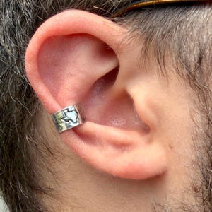 May include: A silver-toned ear cuff featuring a hammered metal texture and a cutout silhouette of the state of Texas. The jewelry piece is worn on the outer cartilage of the ear, showcasing a polished finish and a distinct outline of the state.