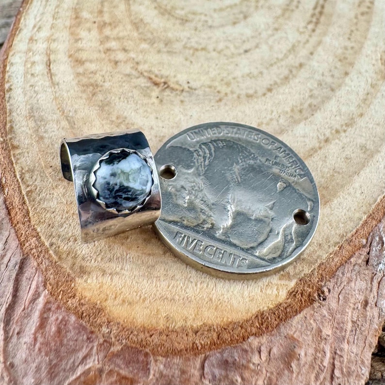 May include: A silver ring with a round, speckled stone in shades of gray and white. The ring is next to a silver coin with the words "FIVE CENTS" and "UNITED STATES OF AMERICA" visible.