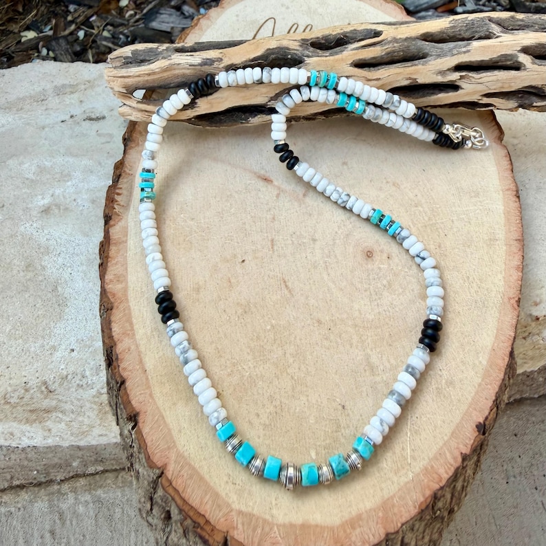 May include: A beaded necklace featuring white, black, and turquoise beads. The necklace has a mix of round and cylindrical beads, with silver-colored spacers. The necklace is displayed on a wooden surface.
