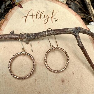 May include: A pair of gold-toned dangle earrings featuring circular hoops with a twisted rope texture. The earrings hang from simple curved ear wires. The text Ally K. is visible in the background, written in a dark brown cursive script.