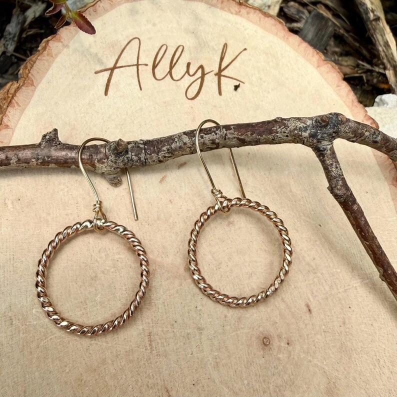 May include: A pair of gold-toned dangle earrings featuring circular hoops with a twisted rope texture. The earrings hang from simple curved ear wires. The text Ally K. is visible in the background, written in a dark brown cursive script.