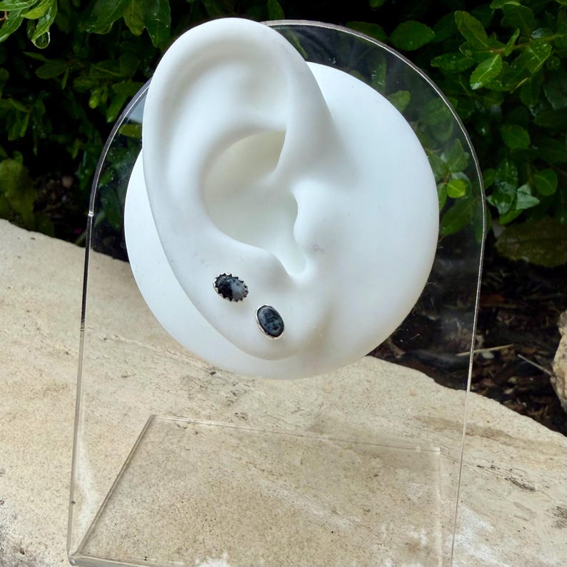 May include: Two stud earrings with dark gray oval and round stones are displayed on a white ear-shaped form. The earrings are set in silver-colored metal. The display is set on a clear acrylic stand.
