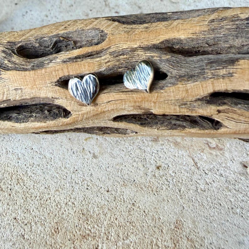 May include: A pair of silver heart-shaped stud earrings featuring a textured, hammered surface. Each earring has a distinct, reflective metallic finish with subtle indentations across the front, creating a light-catching effect on the curved edges.