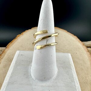 May include: A gold-colored ring with a spiral design. The ring is displayed on a white ring holder against a wooden background. The ring's design features three bands that wrap around the finger.