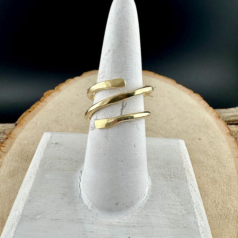 May include: A gold-colored ring with a spiral design. The ring is displayed on a white ring holder against a wooden background. The ring's design features three bands that wrap around the finger.