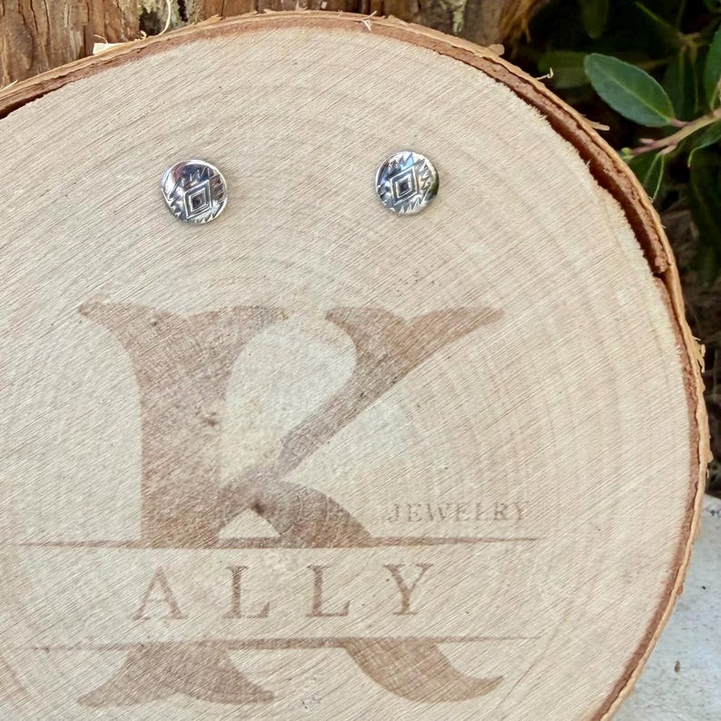 May include: A pair of small, round, silver-toned stud earrings featuring a stamped geometric diamond pattern. The earrings are displayed on a light-colored wooden slice engraved with the text K ALLY JEWELRY.