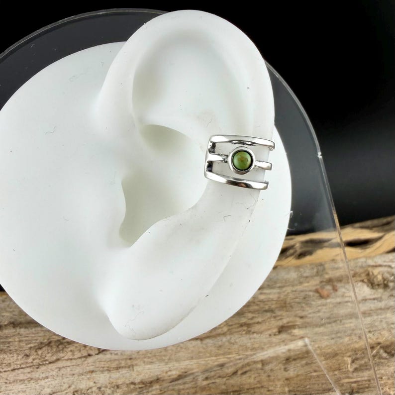 May include: A silver ear cuff with a small, round, green stone set in the center. The cuff has a modern design with three horizontal bars. The jewelry is shown on a white ear model against a neutral background.