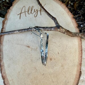 May include: A pair of silver hoop earrings with an etched design. The earrings are hanging from a thin branch, set against a wooden background. The word "Ally" is written in cursive on the wood.