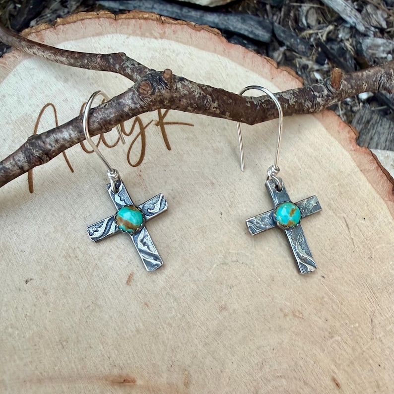 May include: A pair of silver cross earrings with turquoise-colored centers. The crosses have a textured design and are suspended from silver hooks. The earrings are displayed on a wooden surface with a branch.