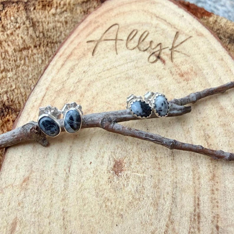 May include: Two pairs of oval-shaped stud earrings with a silver setting and a speckled black and white stone. The earrings are displayed on a small wooden branch, set against a wooden background. The name "Allyk" is carved into the wood.