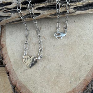May include: A silver-toned necklace featuring a hammered heart-shaped pendant attached to a textured oval link chain. The chain includes a lobster claw clasp closure, with the metal links showing a distinct repeating pattern along their edges.