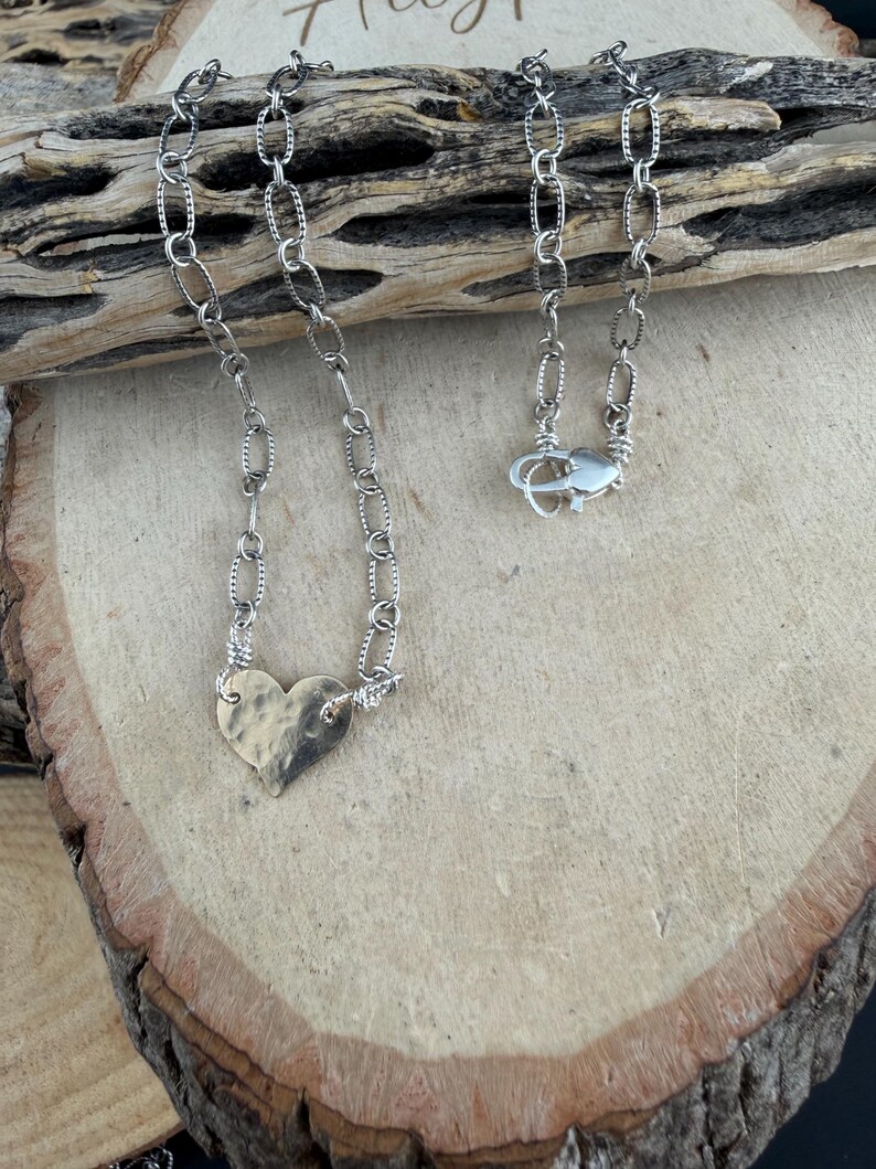 May include: A silver-toned necklace featuring a hammered heart-shaped pendant attached to a textured oval link chain. The chain includes a lobster claw clasp closure, with the metal links showing a distinct repeating pattern along their edges.