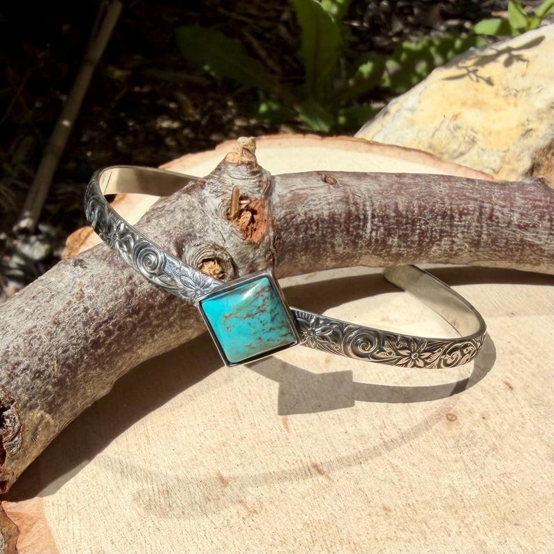 Puede incluir: Una pulsera de plata con una piedra de turquesa cuadrada en el centro. La pulsera presenta grabados florales y en espiral a lo largo de la banda. La turquesa es de un color azul-verdoso vibrante, engastada en un bisel de plata.