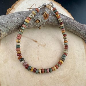 May include: A beaded bracelet featuring a variety of earth-toned beads in shades of red, brown, green, and yellow. The bracelet is strung on a silver chain and is approximately 7 inches in length. The beads are small and round, creating a delicate and colorful accessory.