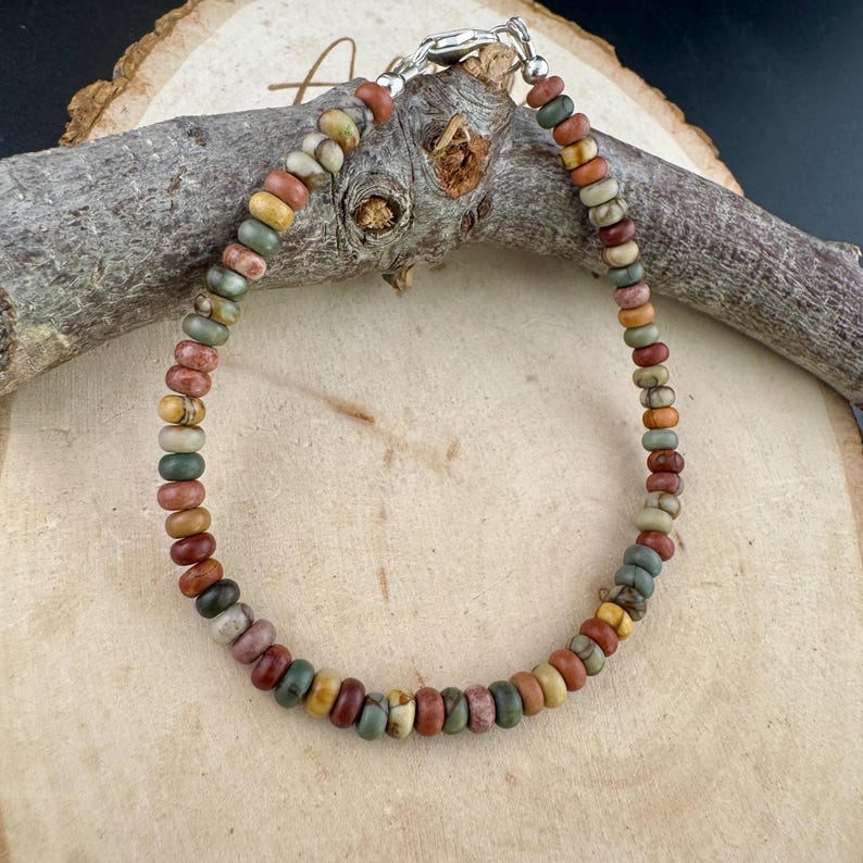 May include: A beaded bracelet featuring a variety of earth-toned beads in shades of red, brown, green, and yellow. The bracelet is strung on a silver chain and is approximately 7 inches in length. The beads are small and round, creating a delicate and colorful accessory.