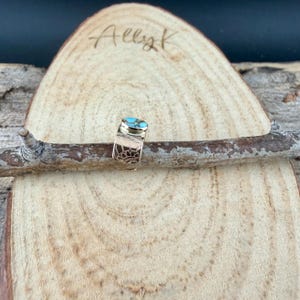 May include: A gold-colored ring with a turquoise stone and a patterned band. The ring is displayed on a wooden surface with the handwritten text "Allyk". The ring's band features a decorative design.
