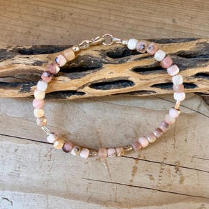 May include: A beaded bracelet featuring small, faceted, square-shaped beads in shades of pale pink, cream, and soft brown. The bracelet is secured with a silver-toned metal lobster claw clasp and includes small silver-toned metal spacer beads.