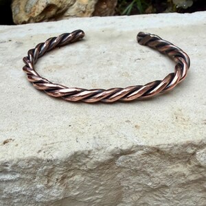 May include: A copper-colored cuff bracelet with a twisted rope design. The bracelet is open-ended and rests on a light-colored stone surface. The metal has a polished finish, reflecting light and showcasing the intricate braiding.