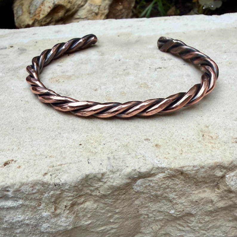 May include: A copper-colored cuff bracelet with a twisted rope design. The bracelet is open-ended and rests on a light-colored stone surface. The metal has a polished finish, reflecting light and showcasing the intricate braiding.