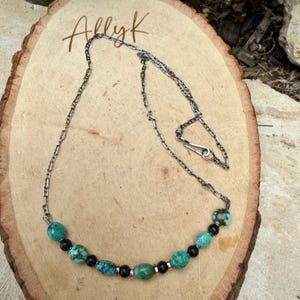 May include: A silver chain necklace with turquoise and black beads. The necklace has a hook clasp and is displayed on a wooden surface. The word "Allyk" is written in cursive on the wood.