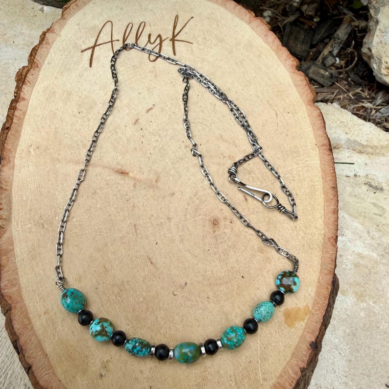 May include: A silver chain necklace with turquoise and black beads. The necklace has a hook clasp and is displayed on a wooden surface. The word "Allyk" is written in cursive on the wood.