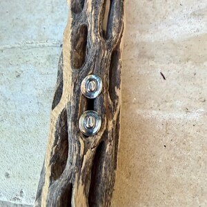May include: Two silver stud earrings with a circular design are displayed on a piece of weathered, light brown wood. The wood has a natural, textured appearance with holes and a rough surface. The earrings are the focal point.