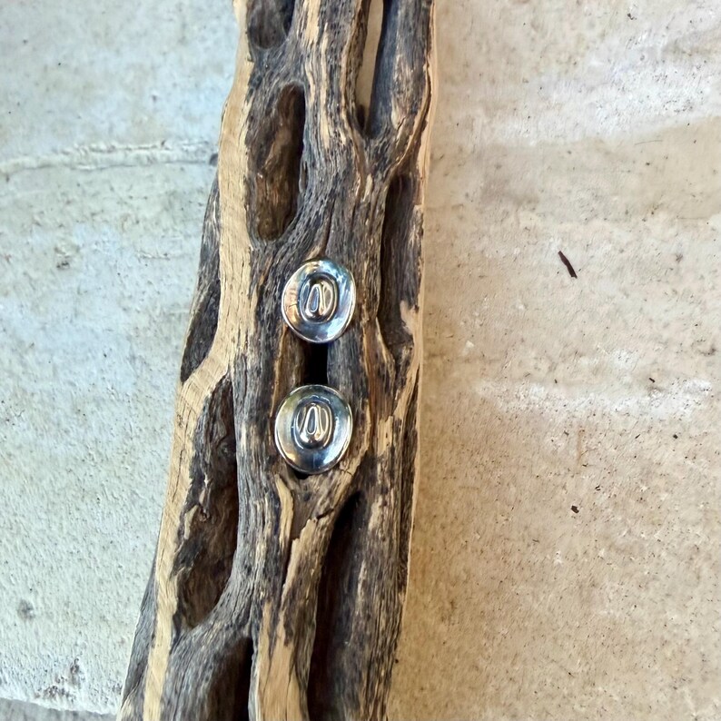 May include: Two silver stud earrings with a circular design are displayed on a piece of weathered, light brown wood. The wood has a natural, textured appearance with holes and a rough surface. The earrings are the focal point.