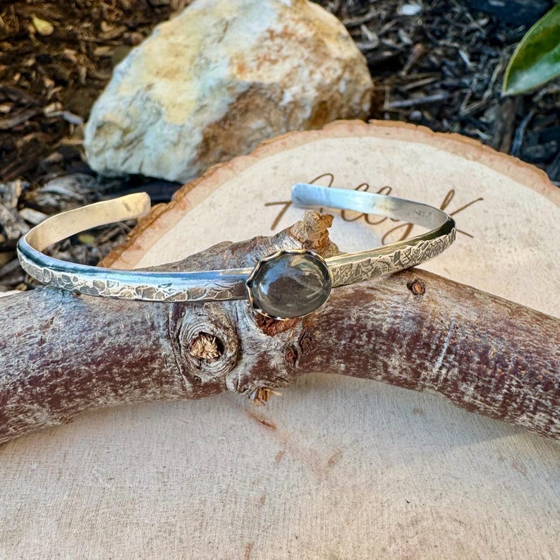 May include: A silver cuff bracelet with a textured band and a dark, oval-shaped stone at the center. The bracelet is displayed on a piece of wood, showcasing its handcrafted details and design.