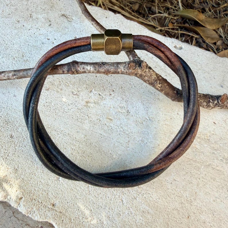 May include: A bracelet composed of two intertwined leather cords, one in a dark brown and the other in a black tone. The cords are secured together by a cylindrical, metallic brass-coloured clasp with a hexagonal centre detail.