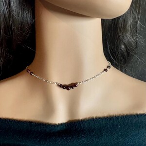 May include: A delicate silver necklace featuring a series of small, dark red beads. The beads are clustered in the center of the necklace, creating a subtle curve. The necklace is designed to sit at the base of the neck, offering a minimalist and elegant look.