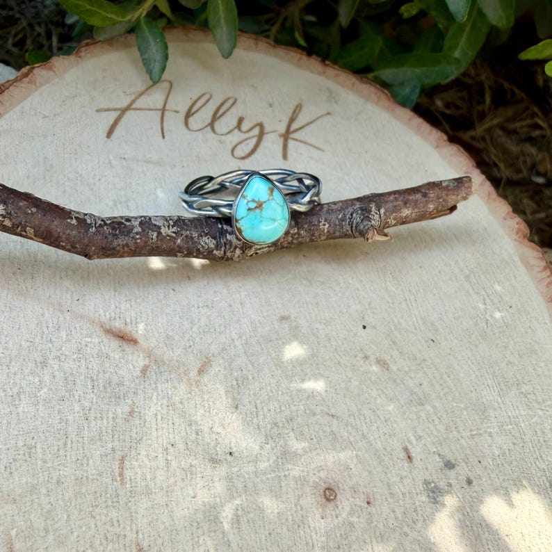 Puede incluir: Un anillo de plata con una piedra turquesa en forma de l&aacute;grima con vetas marrones. La banda est&aacute; hecha de alambre de plata trenzado, creando una apariencia texturizada. El anillo est&aacute; colocado horizontalmente sobre una rama de madera fina.