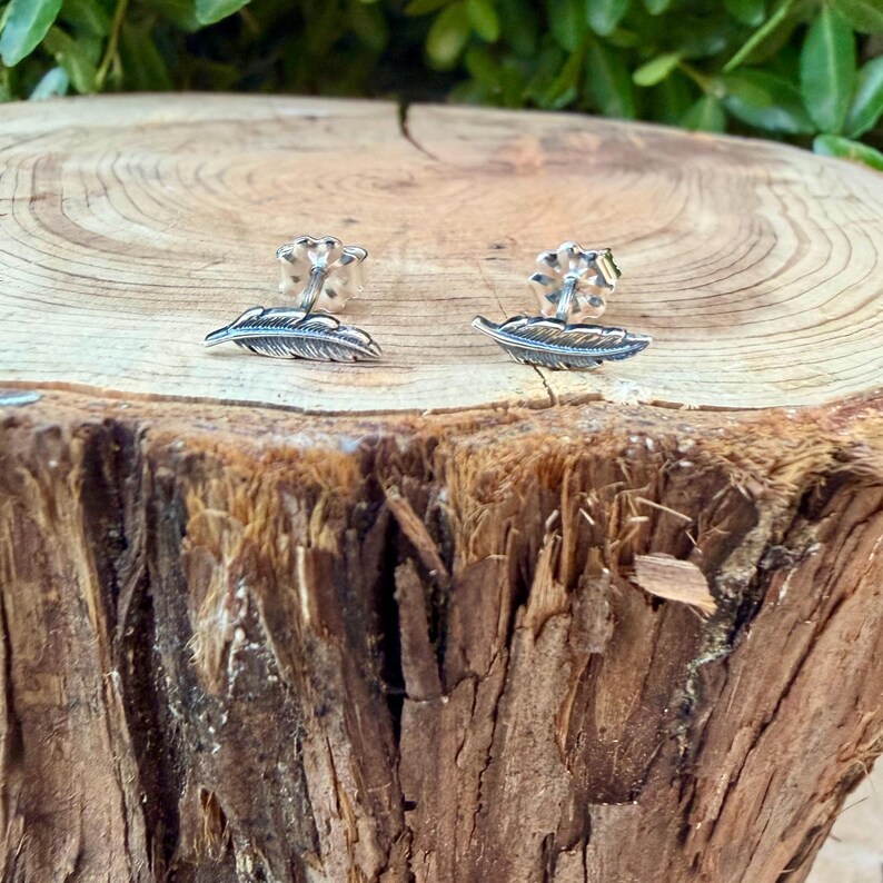 May include: A pair of silver feather stud earrings. Each earring features a detailed feather design with a post backing. The earrings are displayed on a natural wood surface, showcasing their delicate craftsmanship.