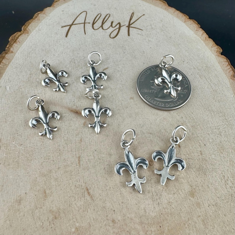 May include: Several small, silver-colored fleur-de-lis charms with attached loops for jewelry making. A U.S. quarter coin is included for size comparison. The charms are displayed on a light-colored, wooden surface. The name "Allyk" is written in cursive.