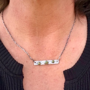 May include: A silver-toned metal bar necklace featuring a textured surface with intricate patterns. The bar is adorned with three small, round, olive-green stones and a central square-shaped olive-green stone, suspended from a delicate chain.