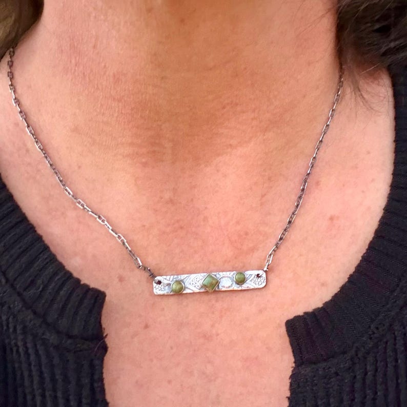 May include: A silver-toned metal bar necklace featuring a textured surface with intricate patterns. The bar is adorned with three small, round, olive-green stones and a central square-shaped olive-green stone, suspended from a delicate chain.