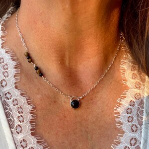 May include: A silver chain necklace featuring a single round black stone pendant at the center and a series of small, alternating brown and black beads integrated into the chain on the left side, resting against a white lace-trimmed garment.