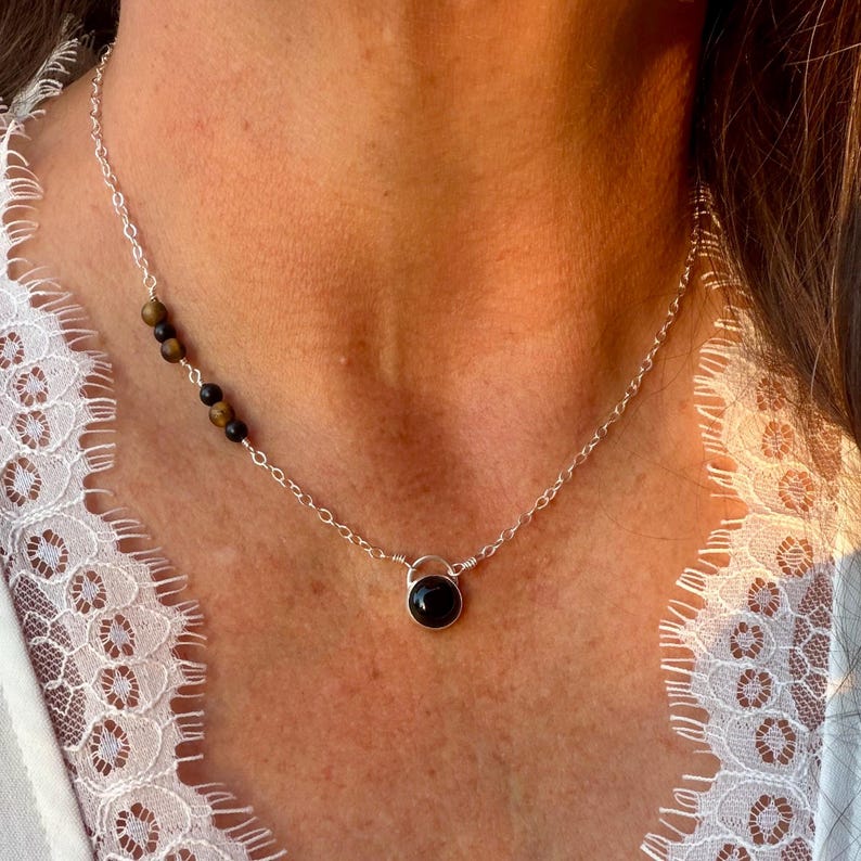 May include: A silver chain necklace featuring a single round black stone pendant at the center and a series of small, alternating brown and black beads integrated into the chain on the left side, resting against a white lace-trimmed garment.