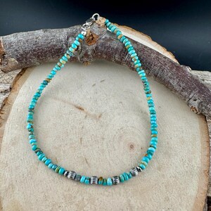 May include: Turquoise beaded bracelet with silver accents. The bracelet features small, round turquoise beads in shades of blue and green, interspersed with silver-colored beads. The bracelet is displayed on a wooden surface.