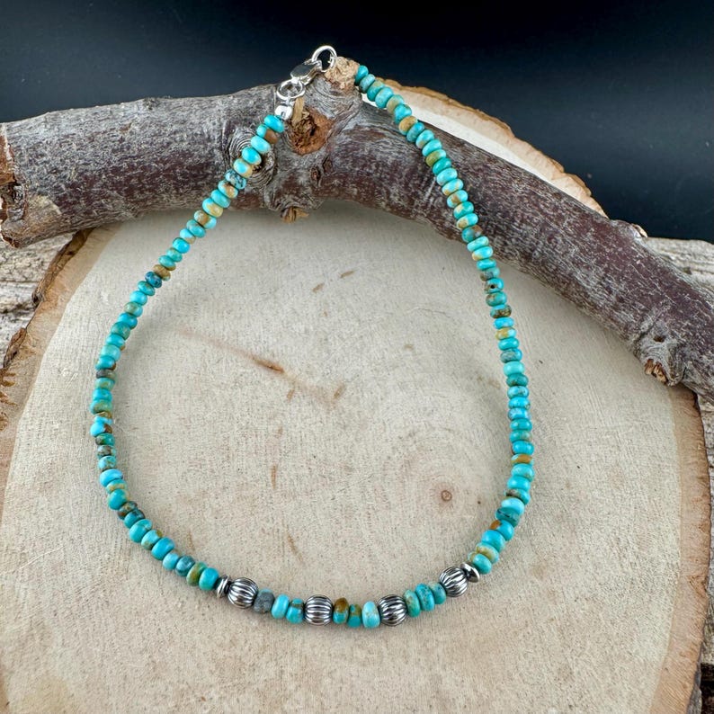 May include: Turquoise beaded bracelet with silver accents. The bracelet features small, round turquoise beads in shades of blue and green, interspersed with silver-colored beads. The bracelet is displayed on a wooden surface.