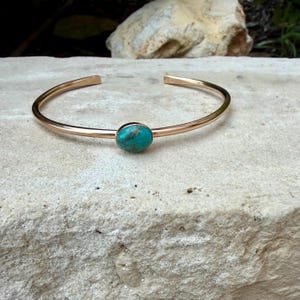 May include: A gold-colored cuff bracelet with an oval turquoise stone set in the center. The bracelet has a simple, open design and is displayed on a textured, light-colored surface.