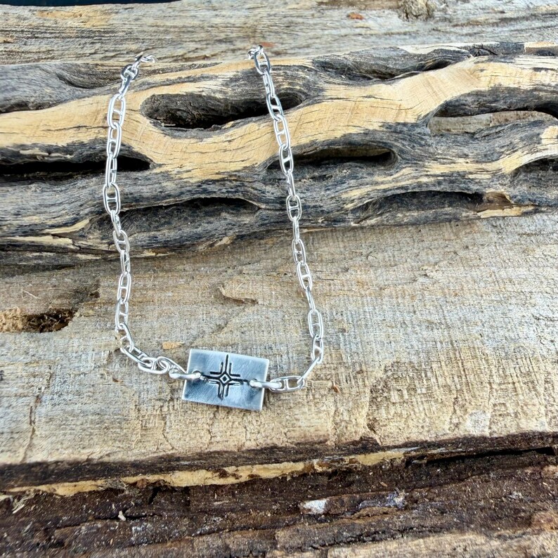May include: A silver-toned necklace with a rectangular pendant featuring a cross design. The necklace has a chain link style and is displayed on a wooden surface. The pendant is approximately 0.75 inches wide.