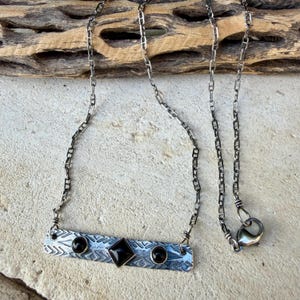 Pu&ograve; includere: Una collana a barra in metallo color argento con un motivo geometrico testurizzato. La barra &egrave; centrata da una pietra nera a forma di diamante, affiancata da due piccole pietre nere rotonde. Il gioiello pende da una sottile catena con moschettone.