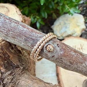May include: Two gold-colored, twisted rope design rings are displayed on a brown wooden branch. The rings are stacked together, showcasing their intricate texture and warm metallic hue. The background features blurred greenery and wood.