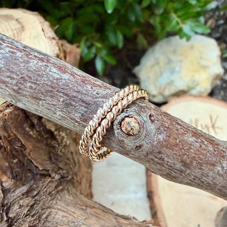 May include: Two gold-colored, twisted rope design rings are displayed on a brown wooden branch. The rings are stacked together, showcasing their intricate texture and warm metallic hue. The background features blurred greenery and wood.