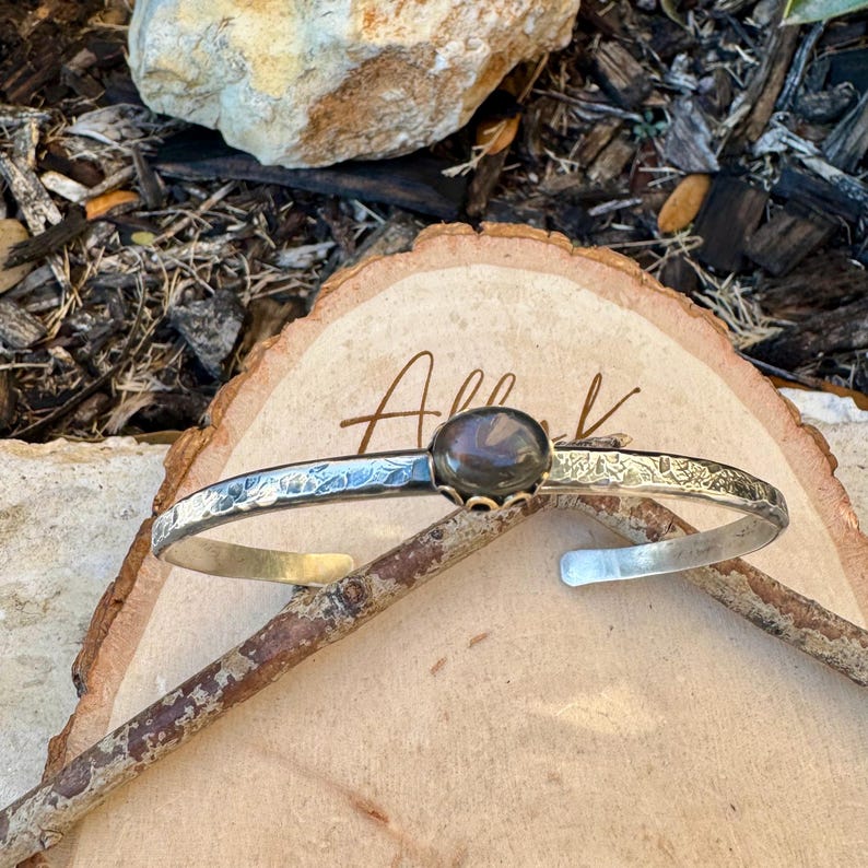 May include: Silver cuff bracelet with a dark, oval-shaped stone at the center. The bracelet has a hammered texture and floral design along the edges. The word "All" is written on the wooden surface.