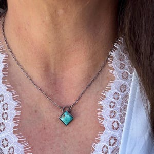 May include: A silver-toned chain necklace with a square turquoise pendant. The pendant is set in a dark metal frame. The necklace is worn against a white lace background.